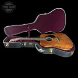 An open acoustic guitar hard case with a wooden acoustic guitar resting inside, photographed against a solid black background. The case has a black exterior with gold-toned latches and a burgundy interior.