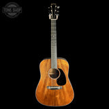 A wooden acoustic guitar with a warm brown finish and black pickguard, photographed upright against a solid black background. A Tone Shop watermark is visible in the top-left corner.