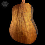 Close-up of the back of an acoustic guitar showing a rich, warm brown wood grain finish, set against a solid black background with a Tone Shop watermark in the top left corner.