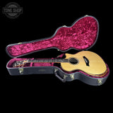 An acoustic guitar with a natural wood body and dark neck, displayed inside an open black hard case with a bright pink velvet interior, set against a solid black background.