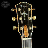 Close-up of a Taylor acoustic guitar headstock with six gold tuning pegs, a dark wood background, and a floral inlay design, shown against a black background with a Tone Shop watermark.