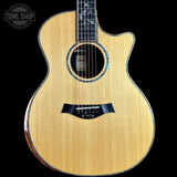 A Taylor acoustic-electric guitar with a natural wood finish and a cutaway body, shown against a solid black background with a Tone Shop watermark in the top-left corner.