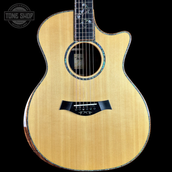 A Taylor acoustic-electric guitar with a natural wood finish and a cutaway body, shown against a solid black background with a Tone Shop watermark in the top-left corner.