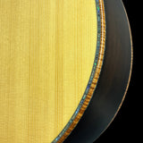 Close-up detail of a guitar body showing the natural wood grain top, a decorative inlay ring, and part of the dark contoured side edge against a black background.