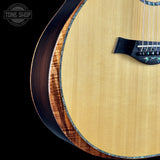 Close-up of an acoustic guitar body showing the natural wood grain top, dark back, and decorative inlay along the edge, set against a black background with a Tone Shop watermark.
