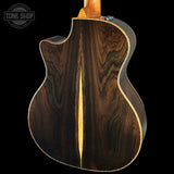 Close-up of the back of an acoustic-electric guitar with a dark brown wood grain body, gold-toned binding, and a light wood neck visible at the top against a black background. A Tone Shop watermark is in the upper left corner.