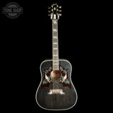 A vintage Gibson acoustic guitar with a dark blackened body, featuring a decorative red, green, and white bird-and-floral inlay on the soundhole surround, set against a solid black background.