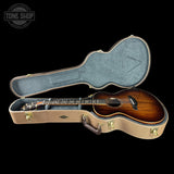 An acoustic guitar with a sunburst finish resting inside an open hard case on a solid black background. The case has a tan exterior and a gray felt interior.