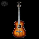 A Taylor acoustic guitar with a natural flame finish, shown vertically against a solid black background. A 'TONE SHOP' watermark is visible in the top-left corner.