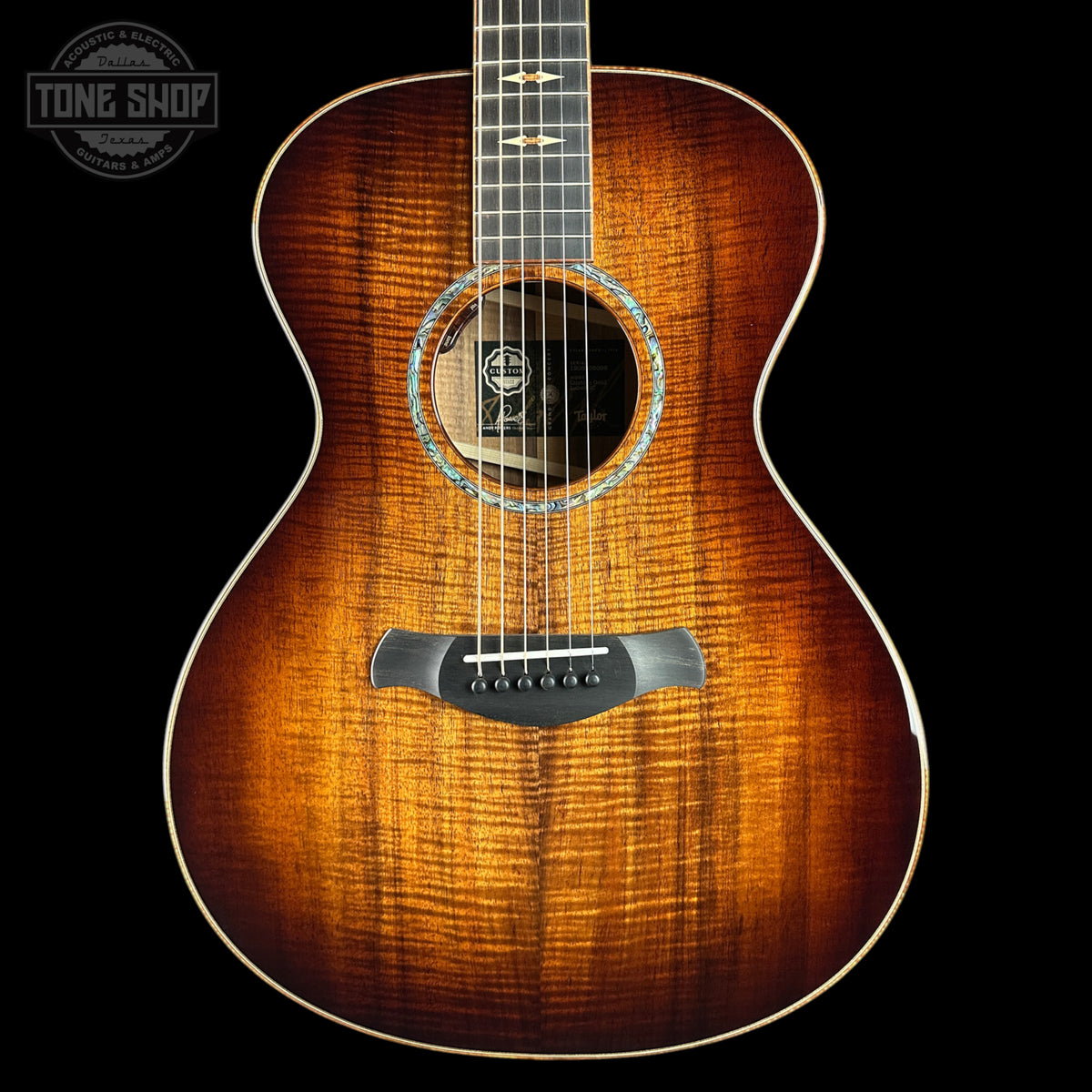 A close-up studio shot of the front of an acoustic guitar against a black background, showing a bright natural wood finish with prominent grain, a black soundhole surround, and a Tone Shop logo overlay in the top left corner.