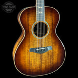 A close-up studio shot of an acoustic guitar body against a black background, showing a warm brown sunburst wood finish with visible grain, a circular soundhole rosette, and part of the neck. A Tone Shop watermark appears in the upper left corner.
