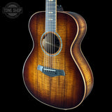 A close-up of an acoustic guitar body with a natural wood finish showing prominent grain, set against a solid black background with a Tone Shop watermark in the top-left corner.