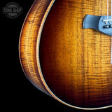 Close-up of an acoustic guitar's flamed wood body showing the rich sunburst finish and grain pattern, with a dark gray bridge on a black background.