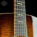 Close-up of an acoustic guitar's neck and body showing a rich brown wood grain finish, dark fretboard, and gold-toned inlays, with a Tone Shop watermark in the top-left corner.
