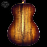 Close-up of the back of an acoustic guitar showing a rich, warm brown wood grain finish on a solid black background, with a Tone Shop logo in the top left corner.
