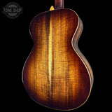 Close-up of the back of an acoustic guitar showing a rich sunburst wood finish with prominent grain patterns, set against a solid black background with a Tone Shop watermark in the top left.