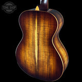 Close-up of the back of a wooden acoustic guitar showing the rich grain and warm brown finish against a black background, with a Tone Shop watermark in the top left corner.