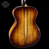 Close-up of the back of an acoustic guitar showing a rich sunburst wood finish with prominent grain patterns, set against a solid black background.