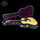 An acoustic guitar with a natural wood body and dark neck displayed inside an open black hard case with a burgundy velvet interior, set against a solid black background. A Tone Shop watermark is visible in the top-left corner.