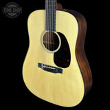 A close-up studio shot of an acoustic guitar against a solid black background, showing the natural wood body, dark pickguard, and part of the neck. A Tone Shop logo is visible in the top-left corner.