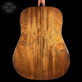 The back of a wooden acoustic guitar, showcasing a rich grain pattern in warm brown and golden tones against a solid black background. A Tone Shop watermark is visible in the top-left corner.
