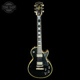 A Gibson Les Paul Custom electric guitar with a black body, gold hardware, and gold trim, shown against a solid black background. A 'TONE SHOP' watermark is visible in the top-left corner.