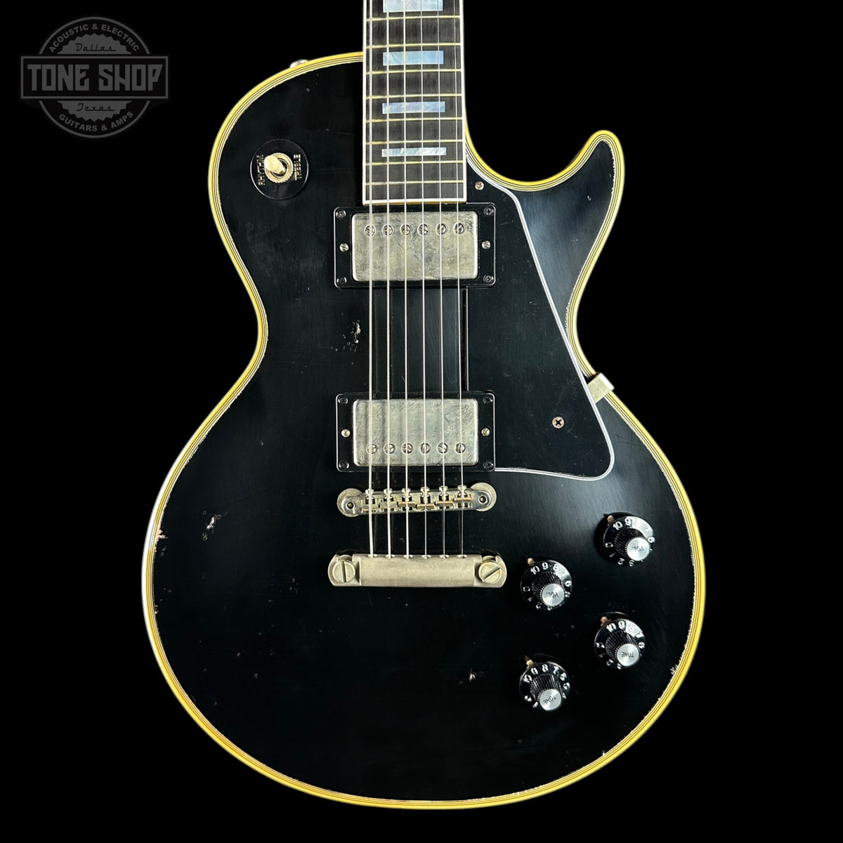 A black electric guitar with a gold outline, featuring a black pickguard, dual humbucker pickups, and four black control knobs, shown against a black background with a Tone Shop logo in the top left corner.