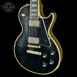 A black electric guitar with gold trim, featuring a double-cutaway body, chrome hardware, and a black pickguard, shown against a solid black background with a Tone Shop watermark in the top left corner.