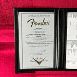 A black leather-bound Fender Certificate of Authenticity document holder, displaying a silver certificate with gold Fender branding on the left and a white receipt sheet on the right, set against a red textured fabric background.