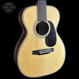 A close-up studio shot of an acoustic guitar body against a solid black background, featuring a natural wood finish, a dark tortoiseshell pickguard, and a white rosette.