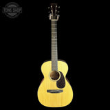 An acoustic guitar with a natural wood finish and black pickguard, shown against a solid black background with a Tone Shop watermark in the top-left corner.