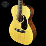A close-up of a wooden acoustic guitar against a solid black background, featuring a natural light-colored spruce top, dark brown sides, and a black pickguard. A Tone Shop logo is visible in the top-left corner.