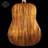 Close-up of the back of an acoustic guitar showing a rich, warm brown wood grain finish, set against a solid black background with a Tone Shop watermark in the top left corner.