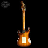 A classic electric guitar shown from the back, featuring a glossy amber-brown sunburst finish, a white pickguard, and gold tuning pegs, set against a solid black background.