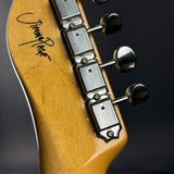 Close-up of the headstock of a Jimmie Dugan electric guitar, showing the maple wood backplate with a black script signature and the silver-and-black tuning pegs against a dark background.