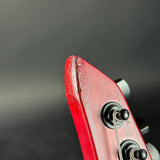 Close-up of the headstock of a red electric guitar, showing the front edge with visible wear and scratches, and the tuning pegs and strings against a dark gray background.