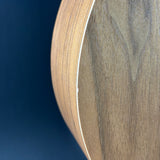 Close-up of a curved wooden object, likely a guitar body or similar instrument component, showing a medium brown wood grain finish and a light binding edge against a dark blue background.