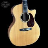 A close-up studio shot of an acoustic guitar against a solid black background, featuring a natural wood finish, a dark tortoiseshell pickguard, and a black bridge. A Tone Shop watermark is visible in the top-left corner.