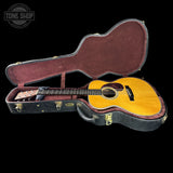 An acoustic guitar with a natural wood top and dark fretboard, displayed inside an open black hard case with burgundy velvet lining, set against a solid black background.