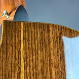 Close-up of a polished wooden instrument body showing a rich grain pattern in warm brown and golden tones, with part of a dark fingerboard and a metal tuning peg visible against a soft gray-blue background.
