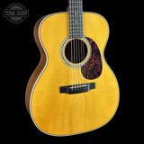 A close-up studio shot of an acoustic guitar body against a solid black background, featuring a natural wood finish, a black pickguard, and a Tone Shop watermark in the upper left corner.