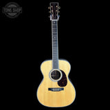 A natural wood acoustic guitar with a dark headstock, shown vertically against a solid black background. A circular 'TONE SHOP' watermark is visible in the top-left corner.