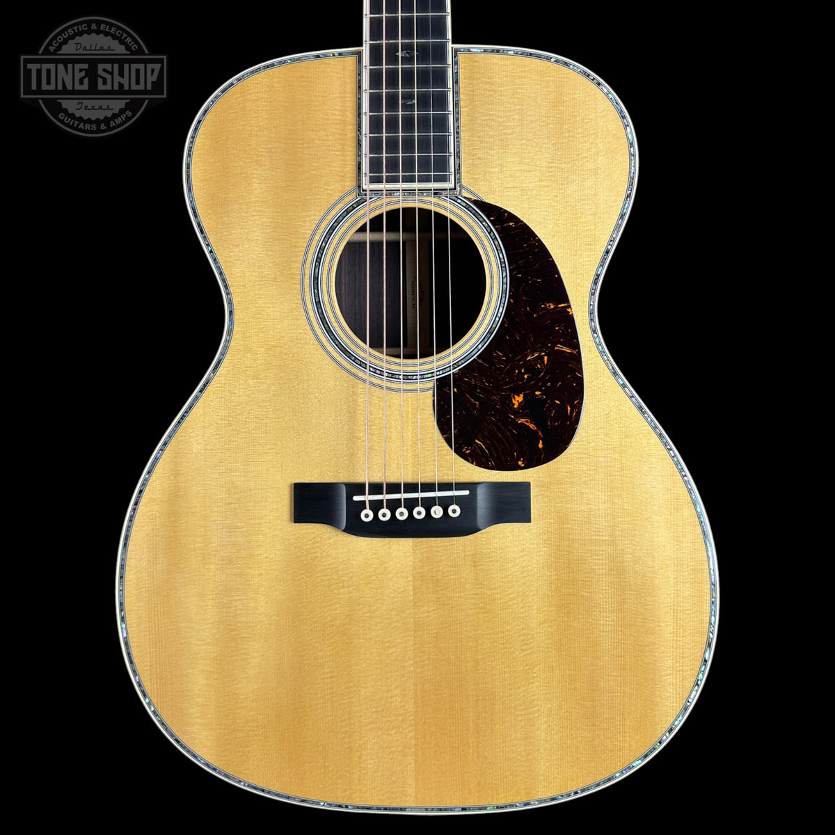 A close-up studio shot of an acoustic guitar body against a black background, showing a natural wood finish, sound hole, pickguard, and bridge. A Tone Shop watermark is visible in the top-left corner.