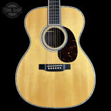A close-up studio shot of an acoustic guitar body against a black background, showing a natural wood finish, sound hole, pickguard, and bridge. A Tone Shop watermark is visible in the top-left corner.