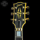 Close-up of a Gibson electric guitar headstock with gold hardware, a black face, and gold logo details, shown against a black background with a Tone Shop watermark in the upper left.
