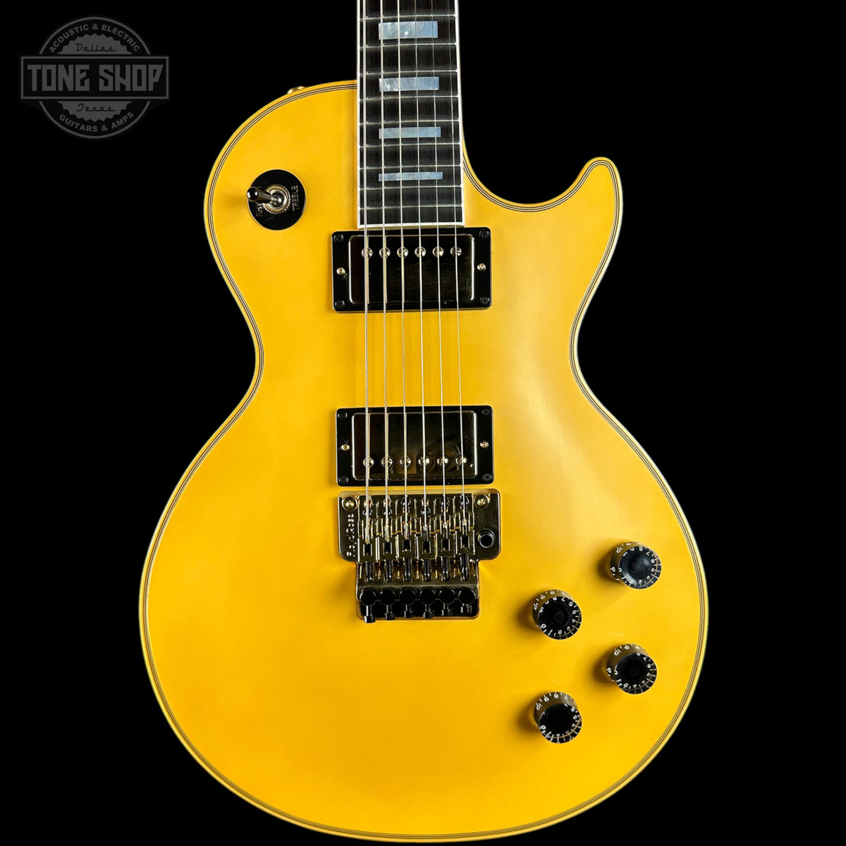 A bright yellow electric guitar with black hardware and control knobs, shown from the front against a solid black background. A Tone Shop logo is visible in the top-left corner.