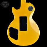 The back of a yellow electric guitar with black pickguard covers and a silver circular badge, shown against a black background with a Tone Shop watermark in the top left.