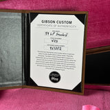 An open Gibson Custom Certificate of Authenticity booklet displayed on a pink textured surface. The white certificate is centered inside a dark brown or black case with a silver-gray interior, and a pink satin ribbon is visible at the bottom right.