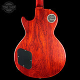 The back of a Gibson Les Paul Custom 'The Magnificent' electric guitar, featuring a vibrant reddish-orange cherry wood finish with a black pickguard and a chrome logo badge, shown against a solid black background.