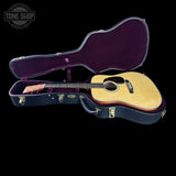 An acoustic guitar with a natural wood body and dark headstock, displayed inside an open black hard case with a purple interior, set against a solid black background. A 'TONE SHOP' watermark is visible in the top left corner.